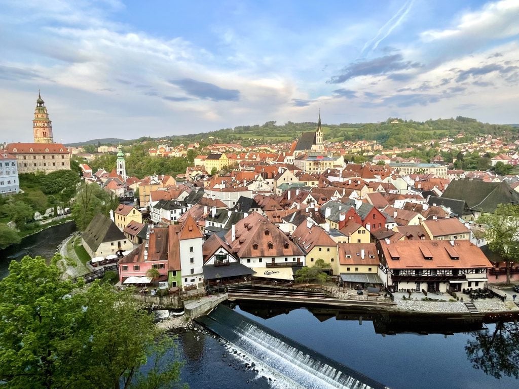 The medieval city of Cesky Krumlov, sitting on a hill, lots of orange-roofed homes and church steeples, all surrounded by a calm river.