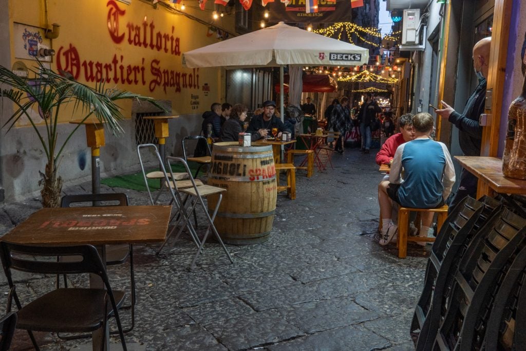 A dark street in Naples, people outside sitting at a dimly lit cafe table.