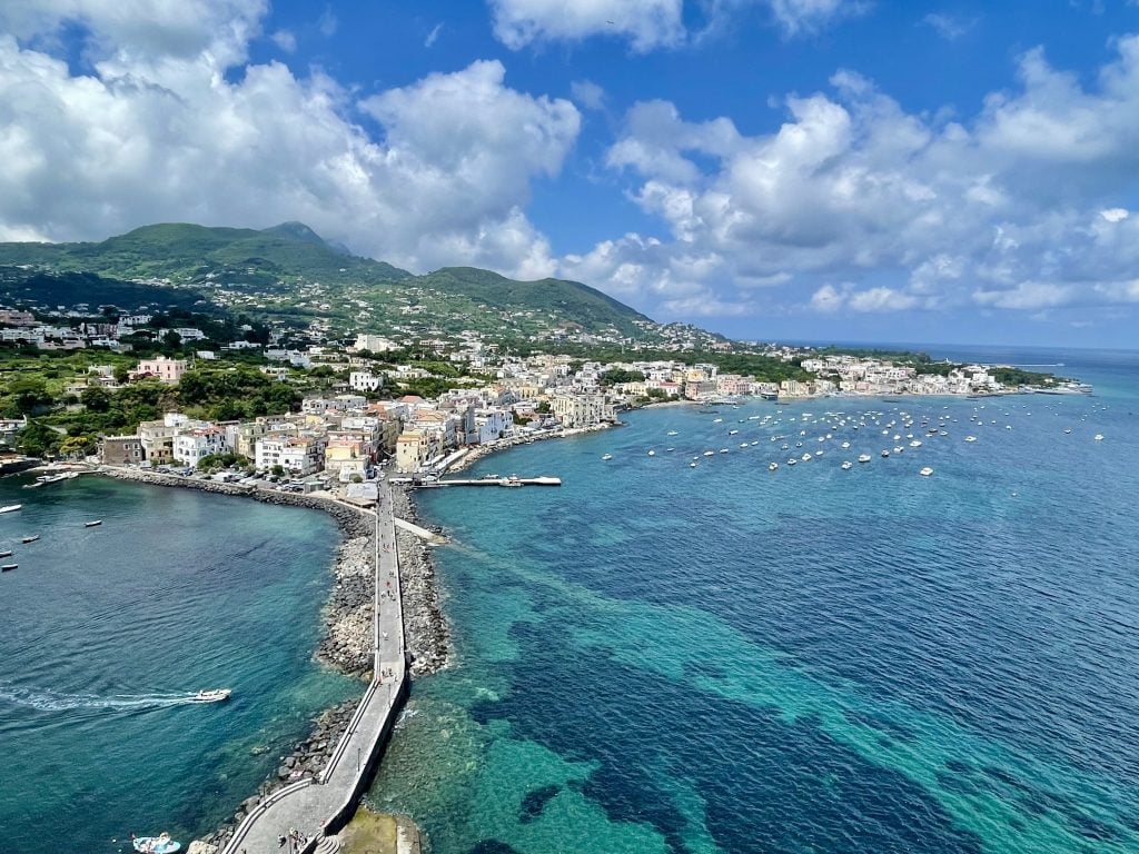 A view from above of a causeway leading to an Italian island covered with white square houses and green mountains. The surrounding water is various shades of turquoise.