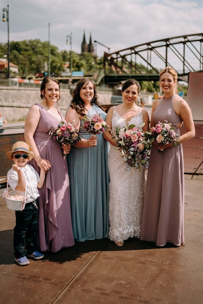 Kate standing with her bridesmaids Beth, Cailin, and Sarah in long gowns in shades of purple and blue. Holding Beth's hand is her five-year-old son in a tiny Panama hat and sunglasses.