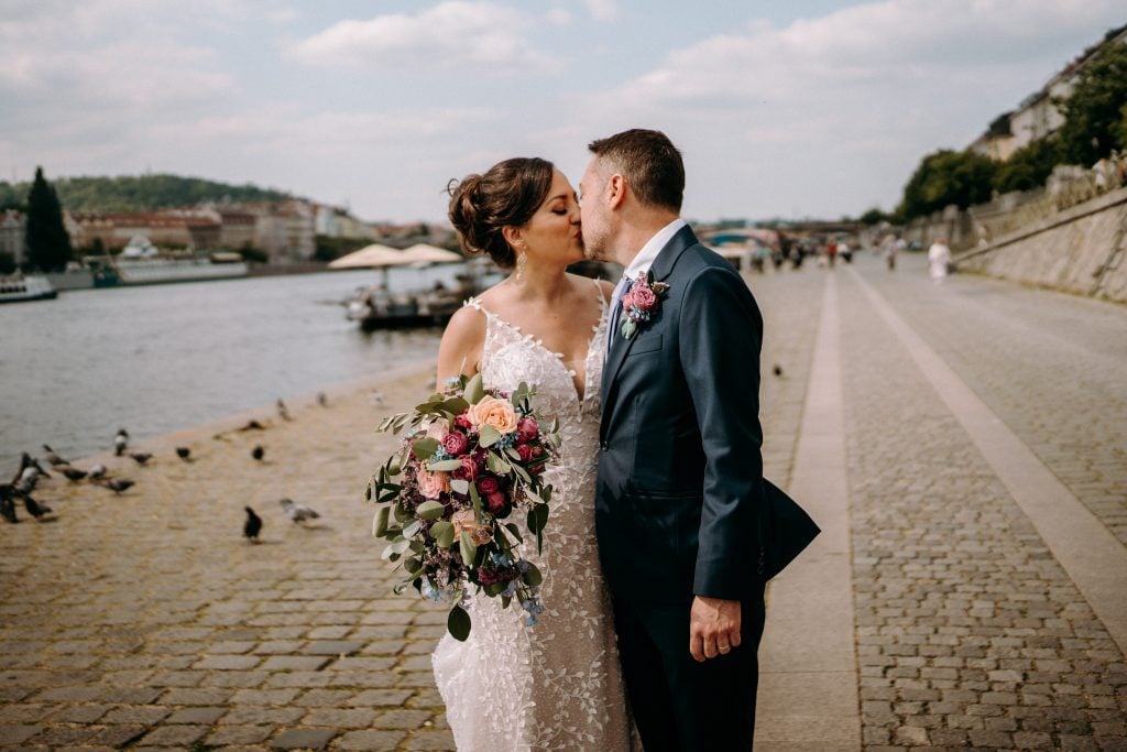 Kate and Charlie kissing on the banks of the river in Prague on their wedding day. Kate wears a white beaded dress with spaghetti straps, has her dark hair in a vintage-style updo, and Charlie wears a navy suit. Kate carries purple, pink, peach, and blue flowers.