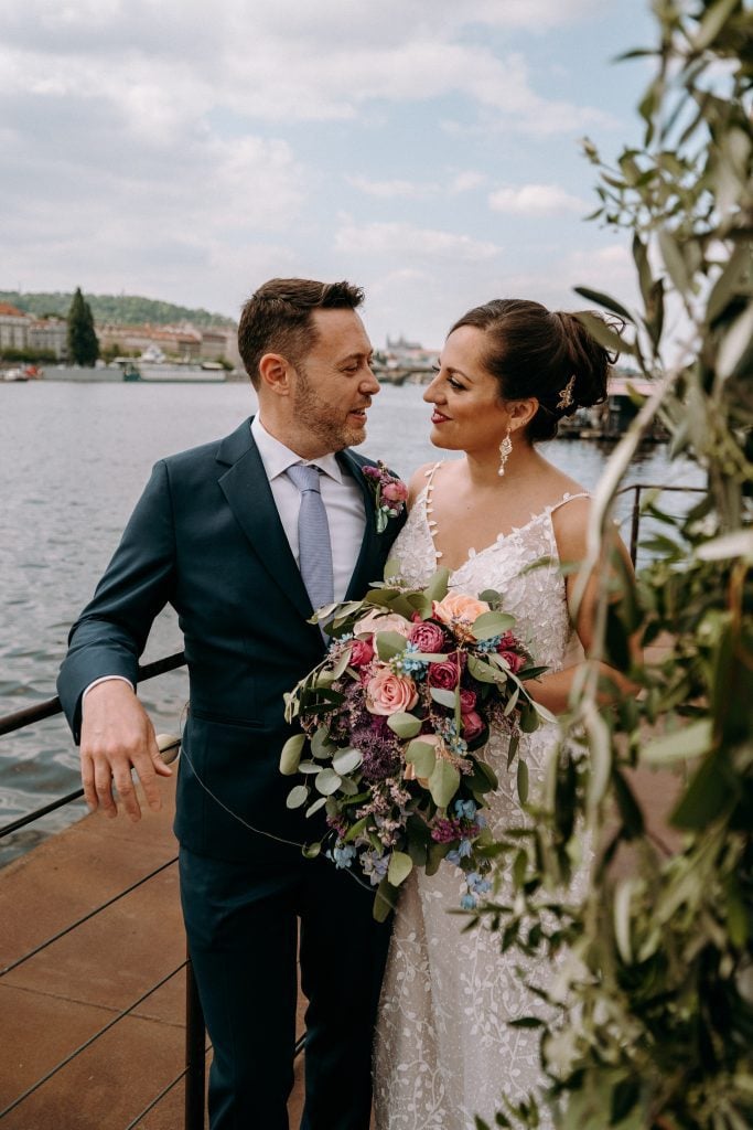 Kate and Charlie smiling at each other on the edge of the boat where they were married. Kate carries a big bouquet of pink, purple, peach and light blue flowers.