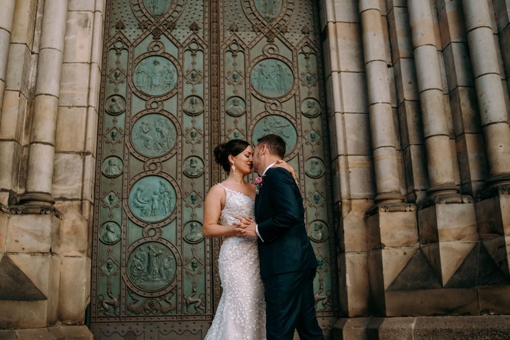 Kate and Charlie in their wedding outfits, embracing in front of a detailed dark green church door.