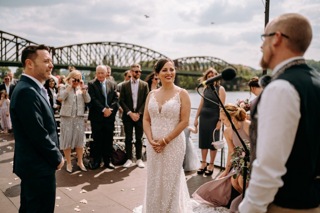 Kate and Charlie on their wedding day (Kate in a long beaded white dress with spaghetti straps) smile at their officiant, wedding guests and a railway bridge behind them.