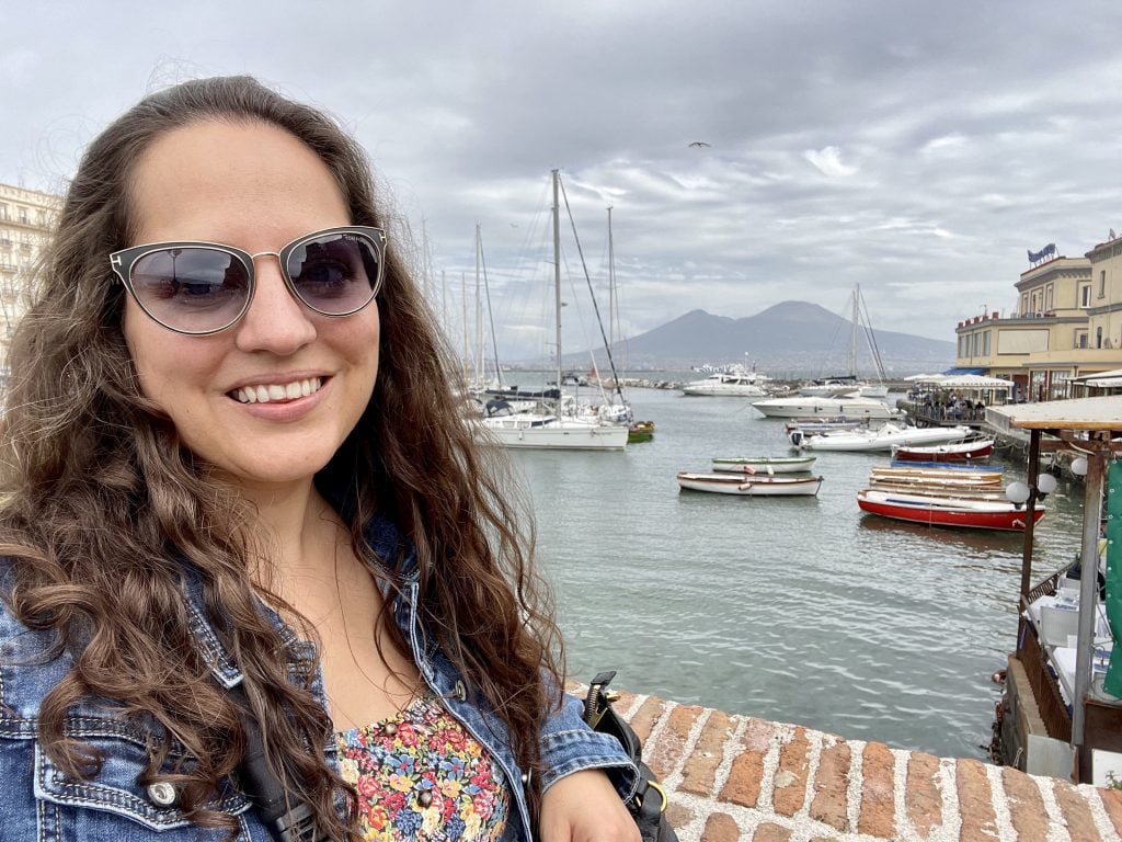 Kate smiling while wearing sunglasses, a denim jacket, and a flowered top. Her hair is long, dark brown, and wavy. Behind her you see a harbor in Naples, with Mount Vesuvius underneath a cloudy sky.