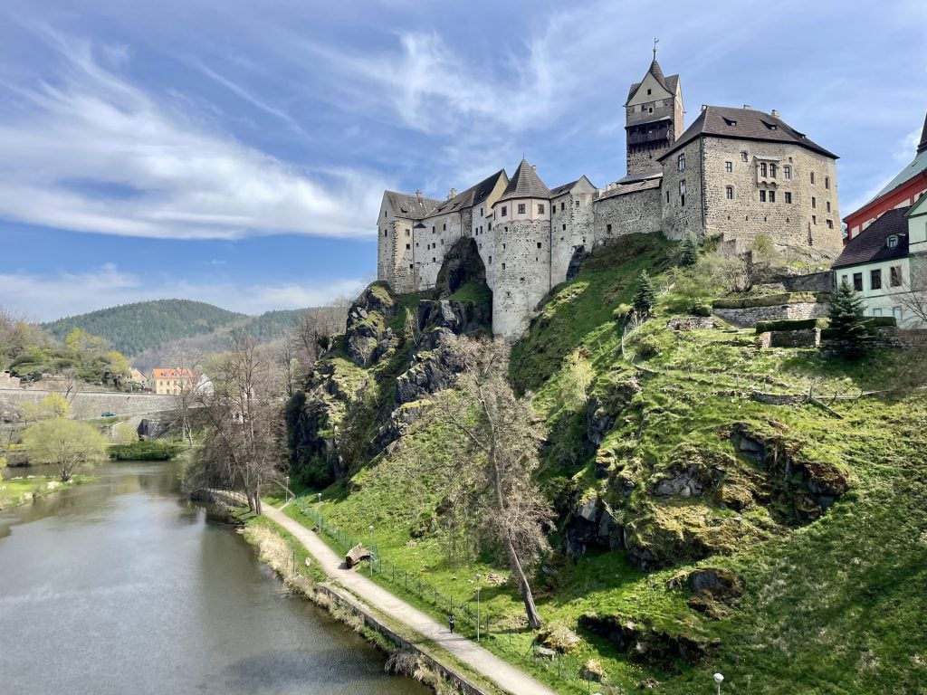 A stone castle built into a craggy hill on the edge of a moat-like river.