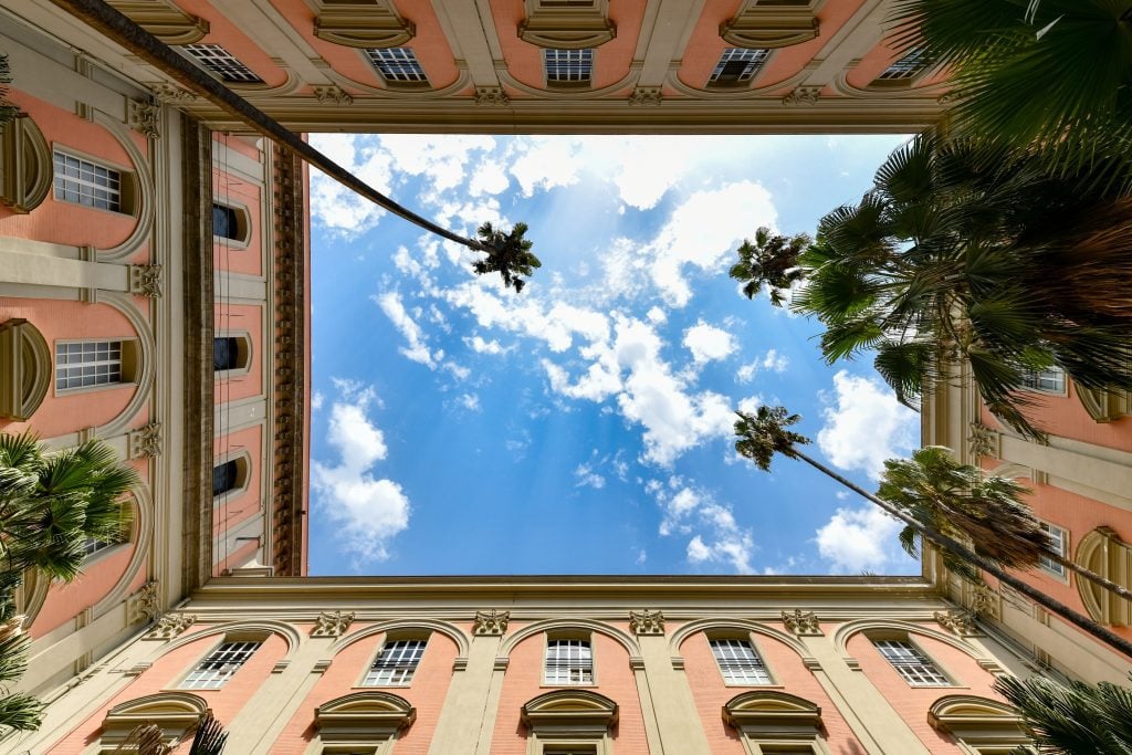 Looking up at the sky from the middle of a courtyard -- you see four pink walls with lots of windows and white panels, and blue sky with clouds in the middle.