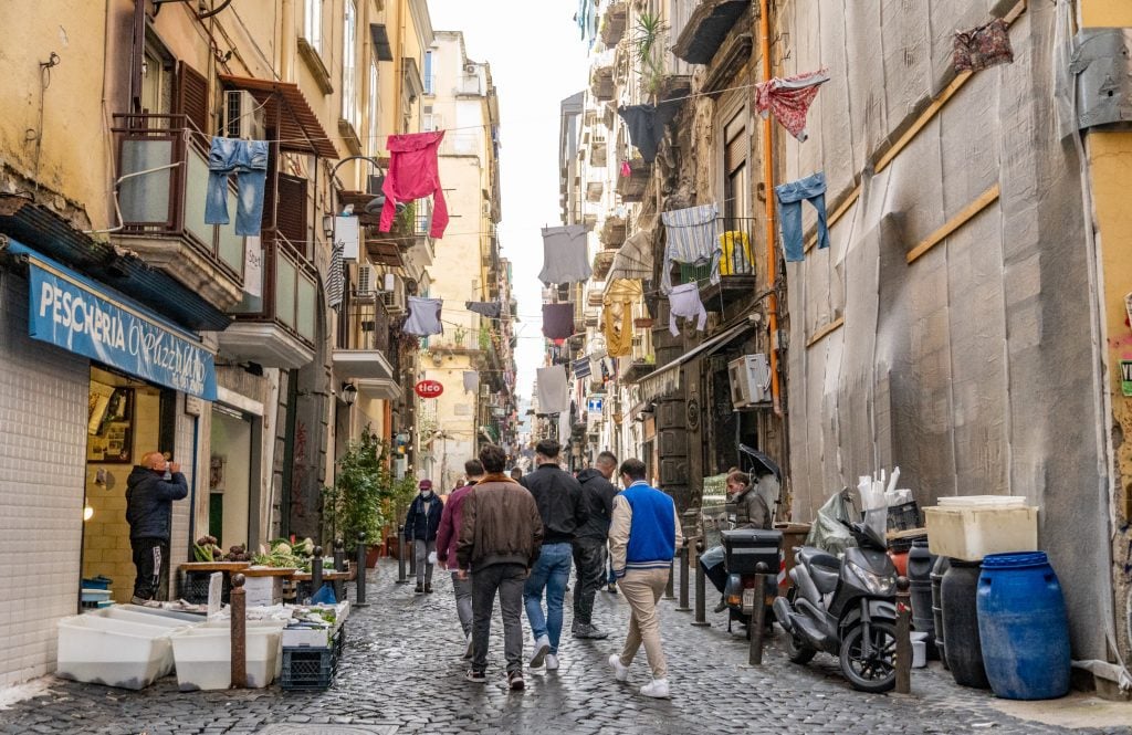Four me walking down a crowded, narrow street in Naples with laundry hanging out the windows and trash bins and cafe tables jockeying for space on the cobblestone street.
