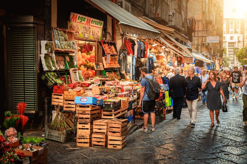 A tourist standing at a fruit and vegetable market on a busy street in Naples.