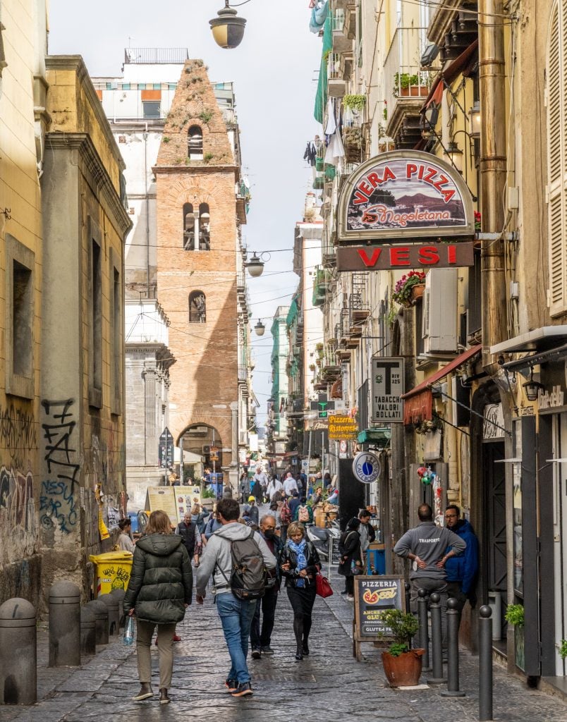A big crowded street in Naples's old town, a church tower in the distance, a neon pizza sign in the foreground!