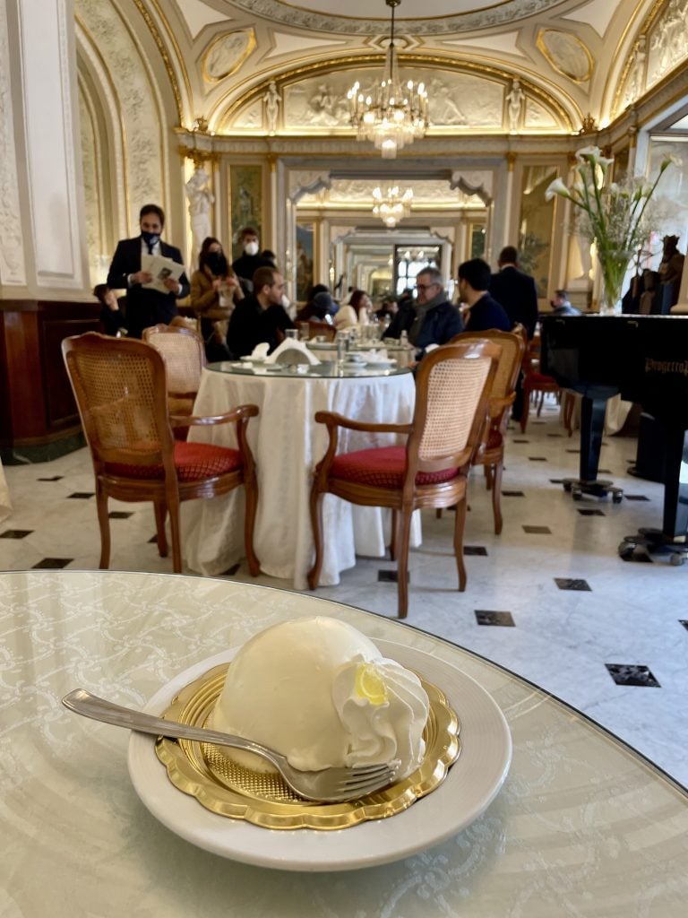 A yellow custard-covered cake on a table in a grand caffe with white and gold crenelated walls.