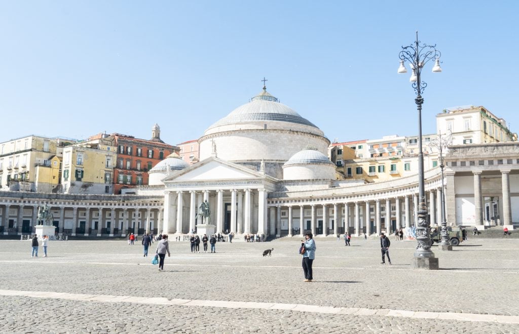 A big white building with a circular dome, surrounding by columns in a colonnade, and a big piazza in front with lots of people walking around.