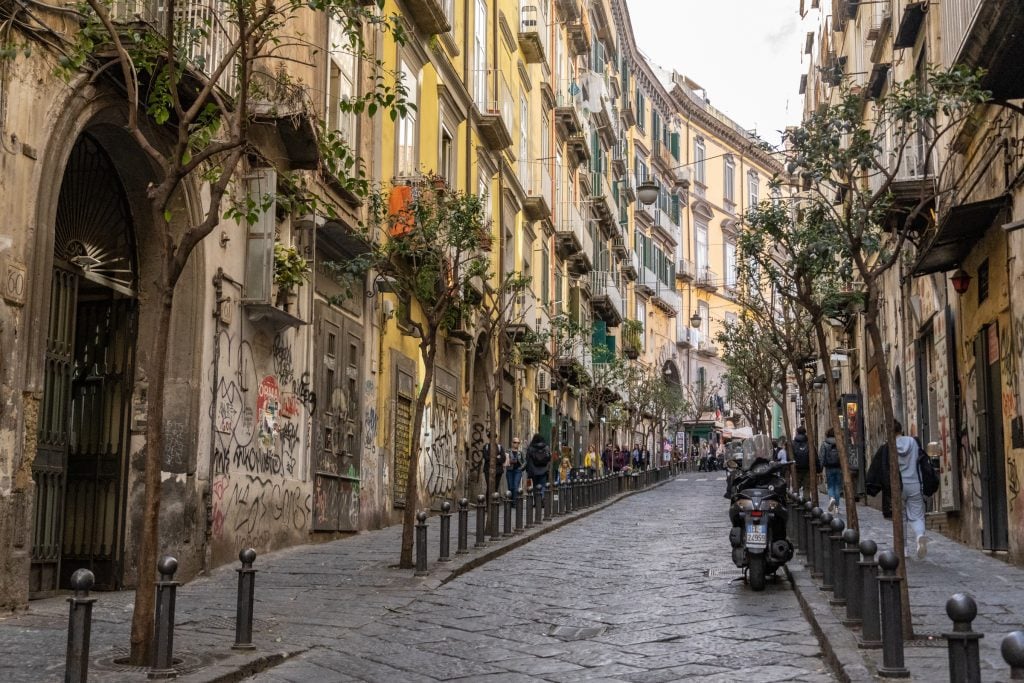 A calm, curved street in Naples, with pale yellow and peach-colored buildings pressed up against each other and scrawny trees lining the street.