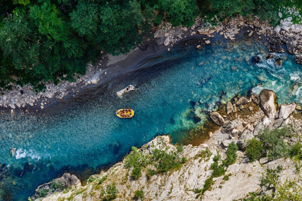 An aerial shot of a big yellow raft in the middle of a bright blue, clear river, surrounded by gray rocks and forest.