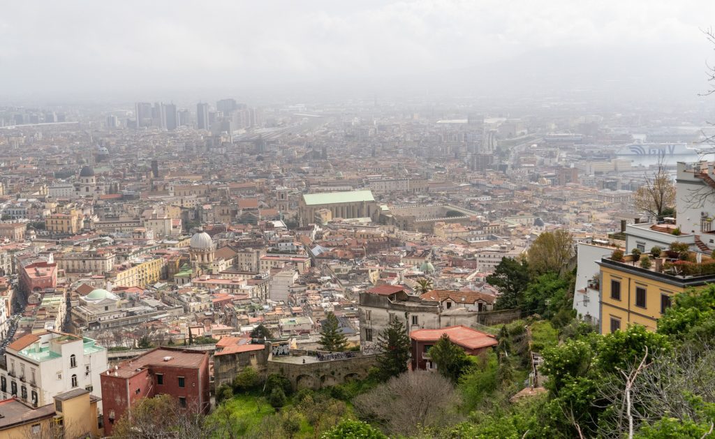 A view from above -- you see a few gardens in the foreground, and behind it, the cityscape of Naples, mostly small buildings, haze hanging overhead.