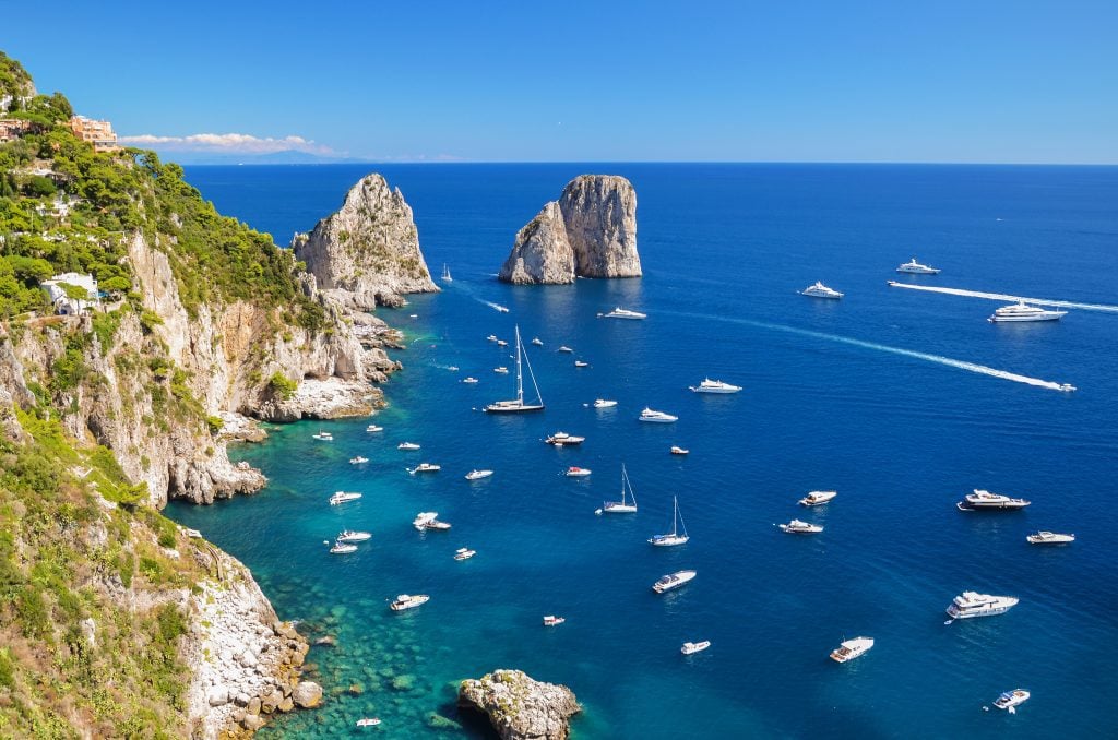 Dozens of small white boats in a calm bright blue bay next to a rocky coastline in Capri.