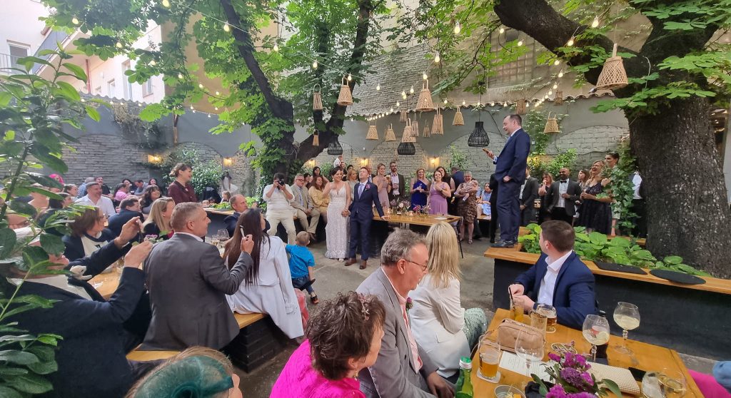 A big greenery filled garden with string lights, lanterns, and crowds of people. The best man stands on a table to make a speech; the bride and groom smile beneath him.