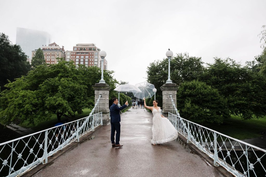 Kate and Charlie in their wedding dress and suit, on the small suspension bridge in the Boston Public Garden. It's a gray rainy day and they both hold clear umbrellas.