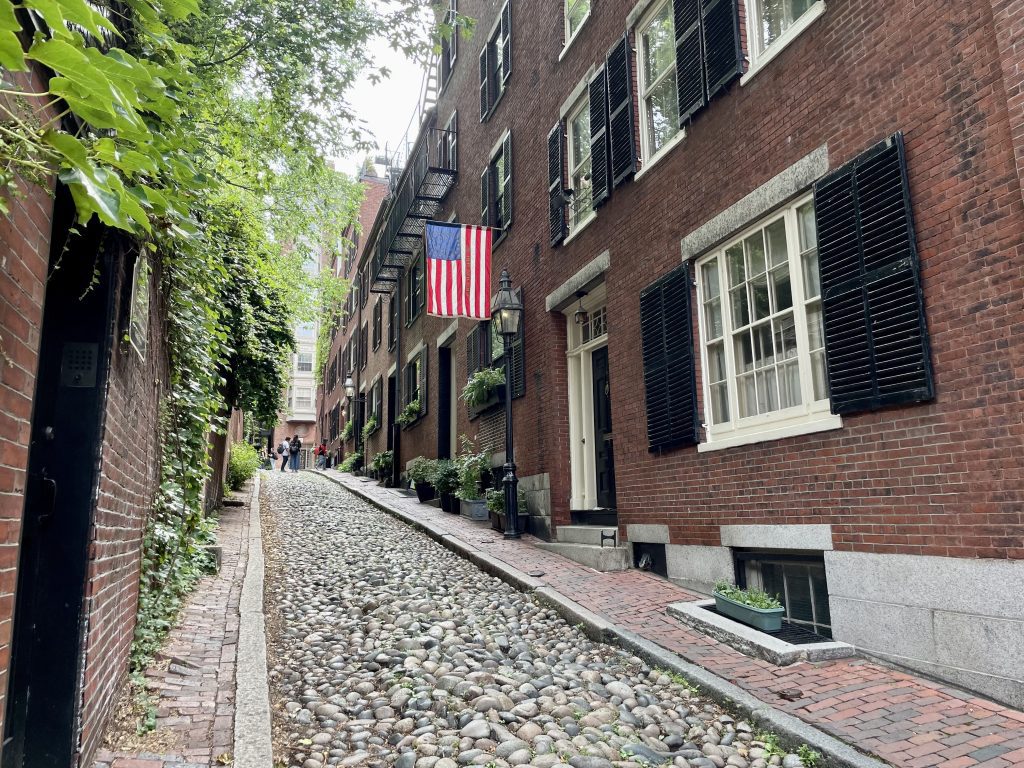A small uphill cobblestone street lined with stately brick buildings with black shutters, one hanging an old-fashioned American flag from the 19th century.