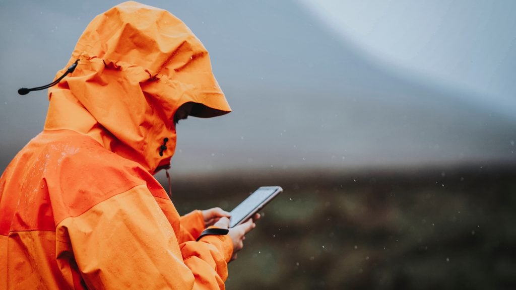 A woman in a hooded orange rain jacket, leaning down to look at her phone.