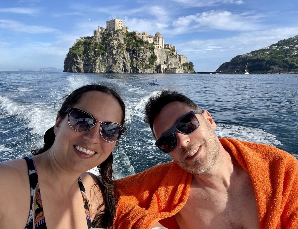 Kate and Charlie taking a smiling selfie on a boat, a castle built on a rock behind them, looking like it's rising out of the sea.