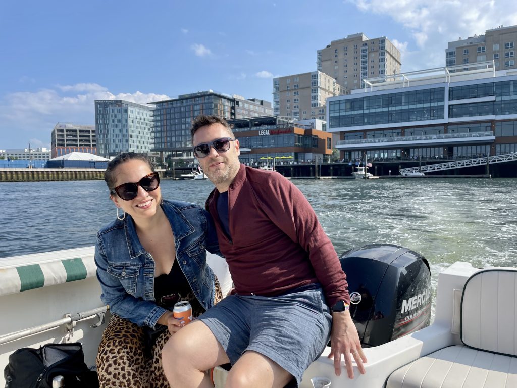 Kate and Charlie riding a small white boat in Boston, posing in front of the harbor with Legal Sea Foods in the background.