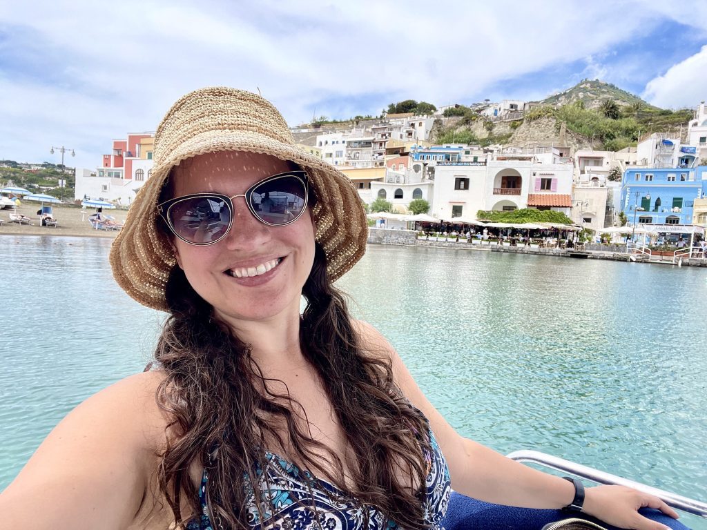 Kate smiling, wearing a straw bucket hat and sunglasses, in front of a small coastal town in Ischia, Italy.