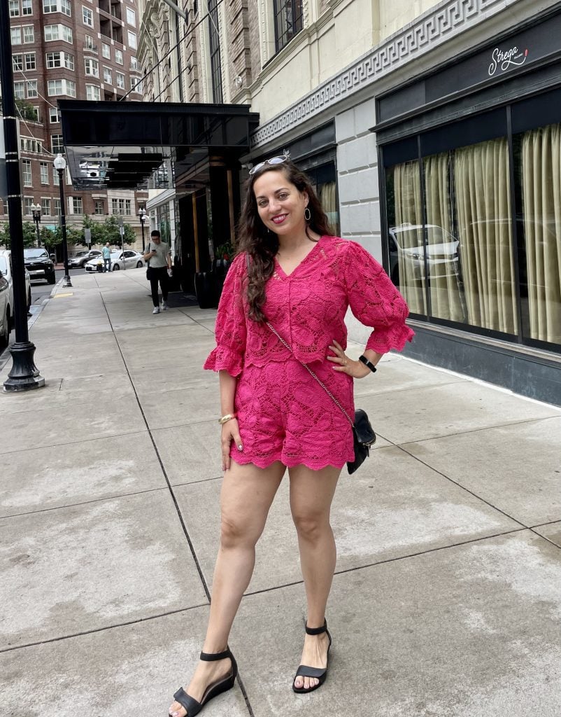 Kate wearing a hot pink outfit: a matching top and shorts in hot pink. The top has elbow-length voluminous sleeves, the shorts are short, and both have a paisley cutout pattern in the fabric.