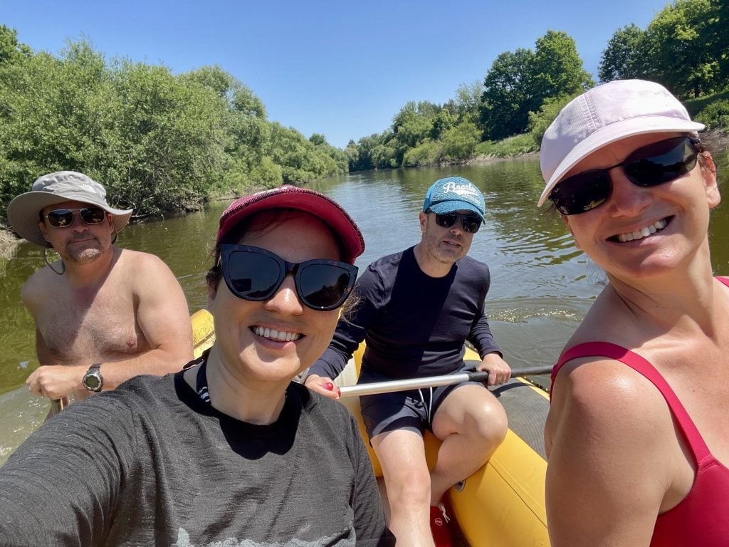 Kate and three friends rafting in a big yellow raft with oars. They all wear hats and sunglasses to ward off the sun.