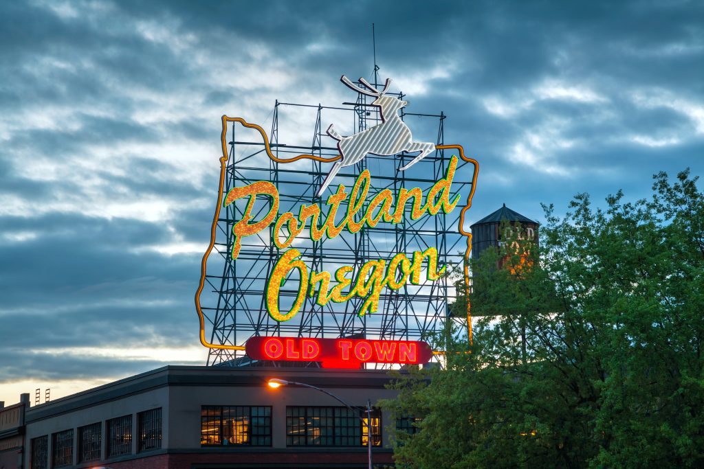 The bright Portland Oregon sign illuminated against the sky, in the shape of the state of Oregon with a jumping deer on top. 