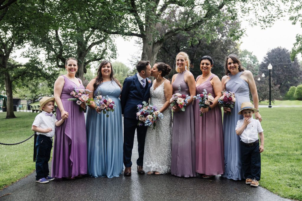 Kate and Charlie in their wedding outfits kissing, surrounded by five girls in light purple and blue dresses, and two little boys in white shirts and Panama hats.