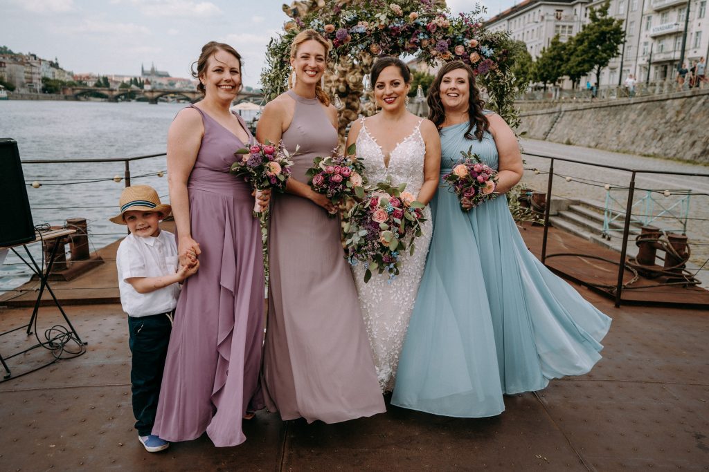 Kate on her Prague wedding day surrounded by three bridesmaids in purple and blue dresses, and a five-year-old boy in a Panama hat holding his bridesmaid mom's hand. 