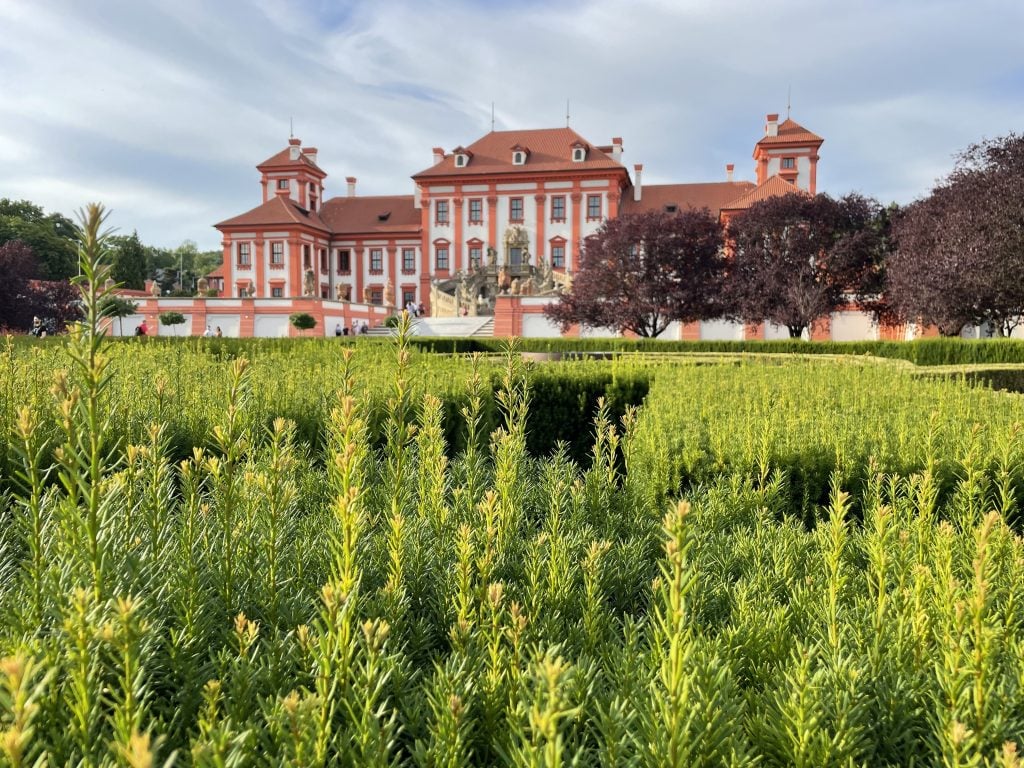 A stately Czech palace in red and white, surrounded by tall green plants.