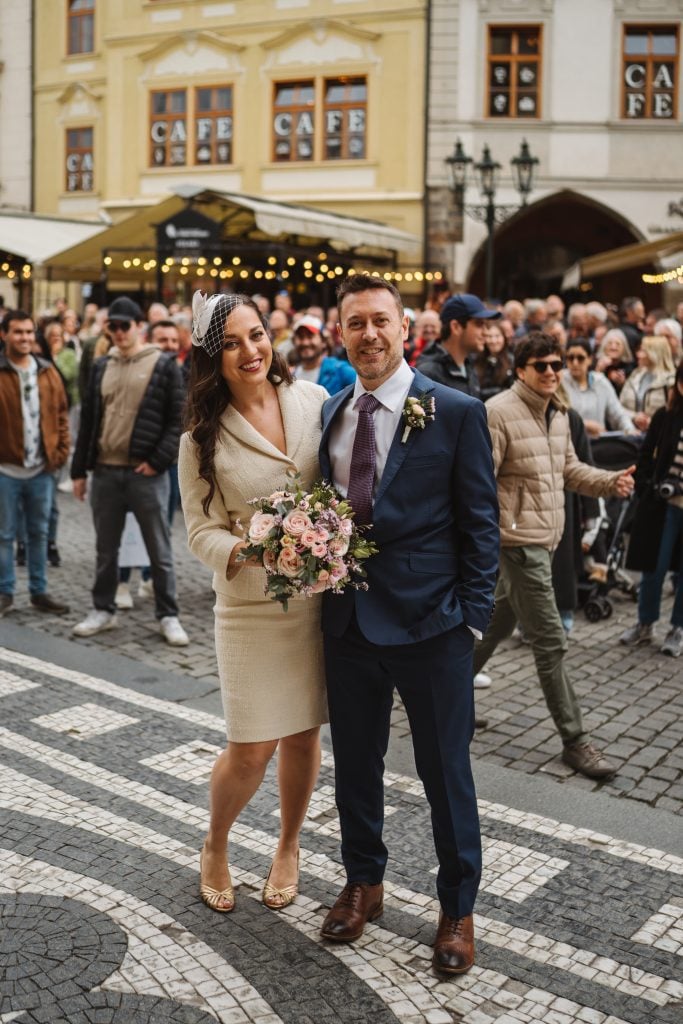 Kate and Charlie at their elopement in Prague, Kate in an ivory skirt suit with a bouquet and Charlie in a navy suit, people walking past behind them.