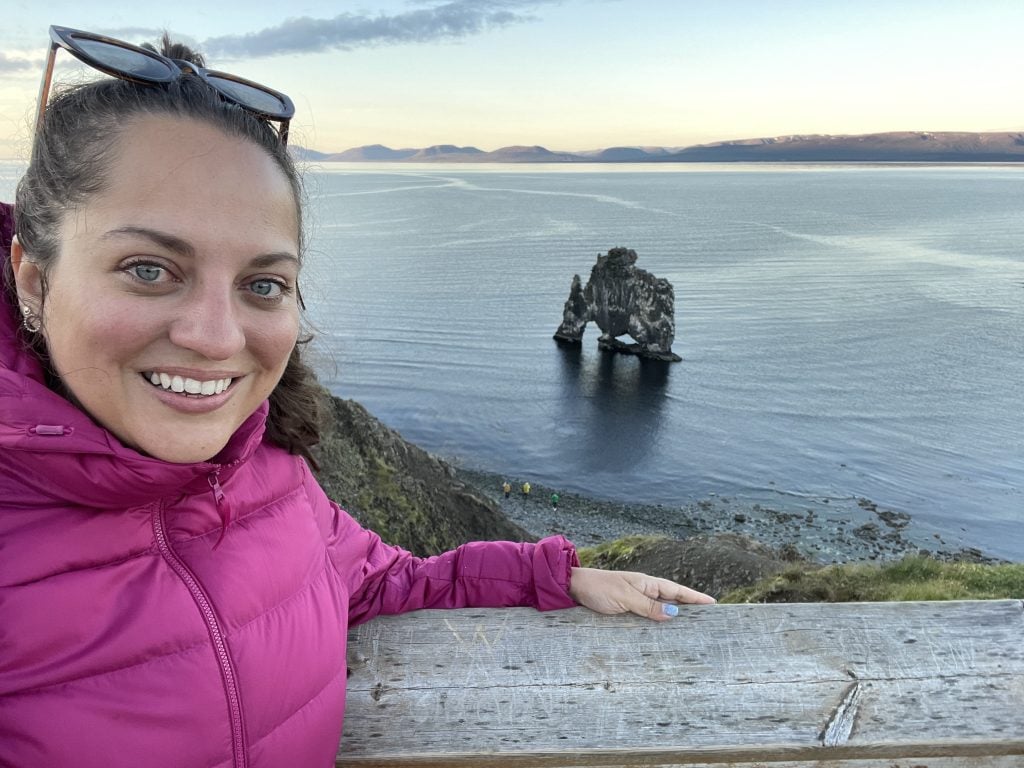 Kate in a bright pink coat taking a selfie from a cliff above Hvitserkur rock in Iceland, which is shaped like a rhino drinking out of the water.