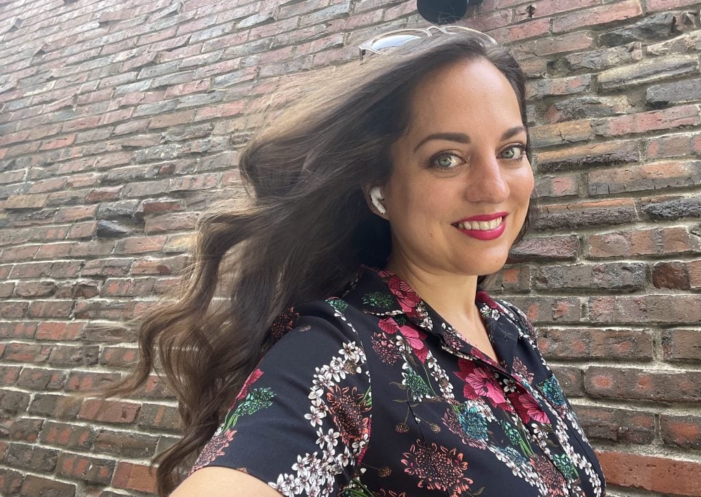 Kate smiling with her long wavy hair flowing behind her as she poses in front of a brick wall.