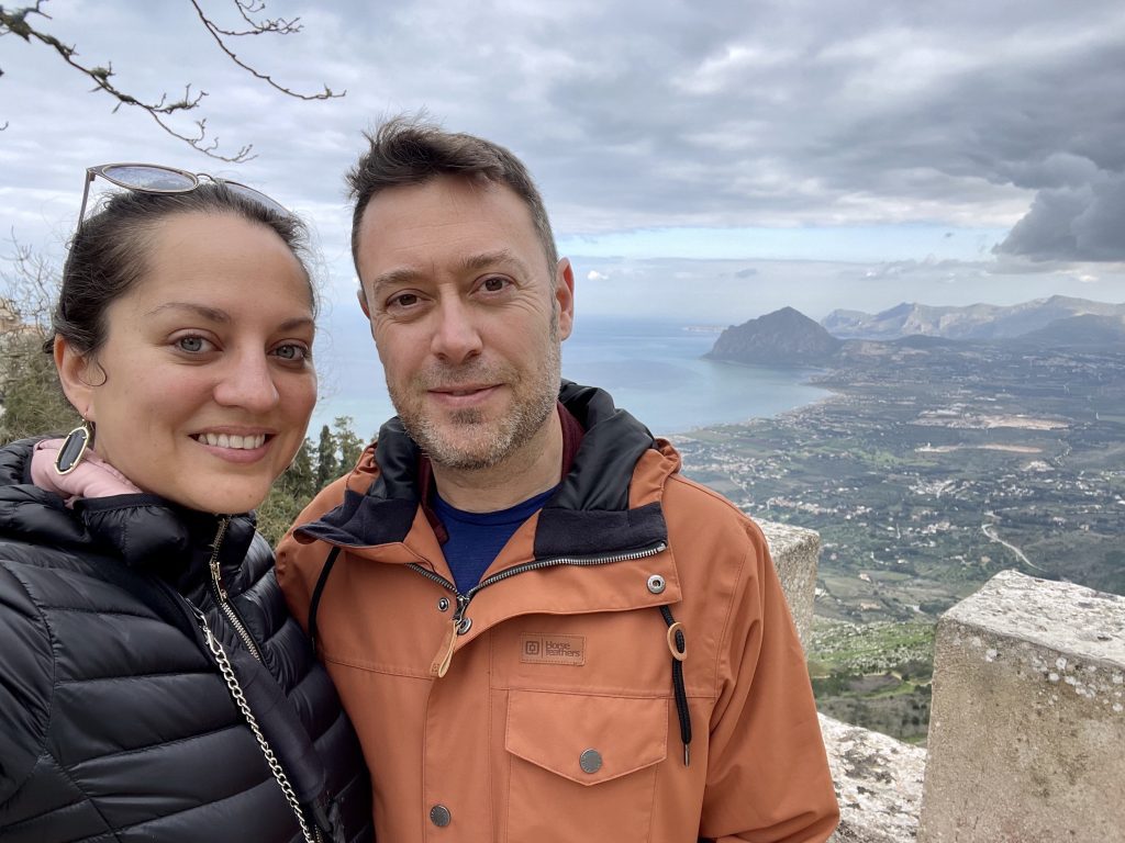 Kate and Charlie taking a selfie from a medieval wall in Erice, Sicily, the landscape splaying out into the distance, mountains and coastline.