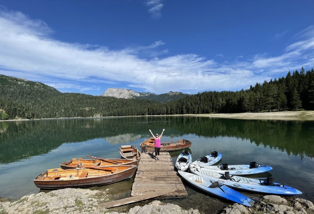 Kate standing on a wooden dock on a still lake reflecting mountains, surrounded by wooden boats and bright kayaks.