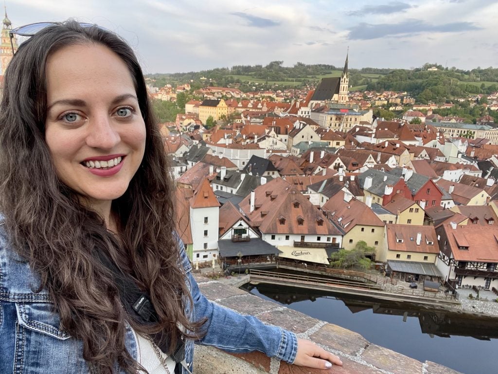 Kate taking a smiling selfie in front of Cesky Krumlov, a medieval city along a small, calm river with a few church towers poking up above the orange-roofed white buildings.