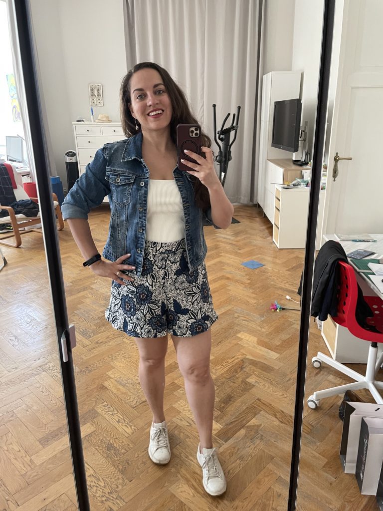 Kate taking a mirror selfie in a pair of white and blue embroidered brocade shorts, a white tank top and blue jean jacket.