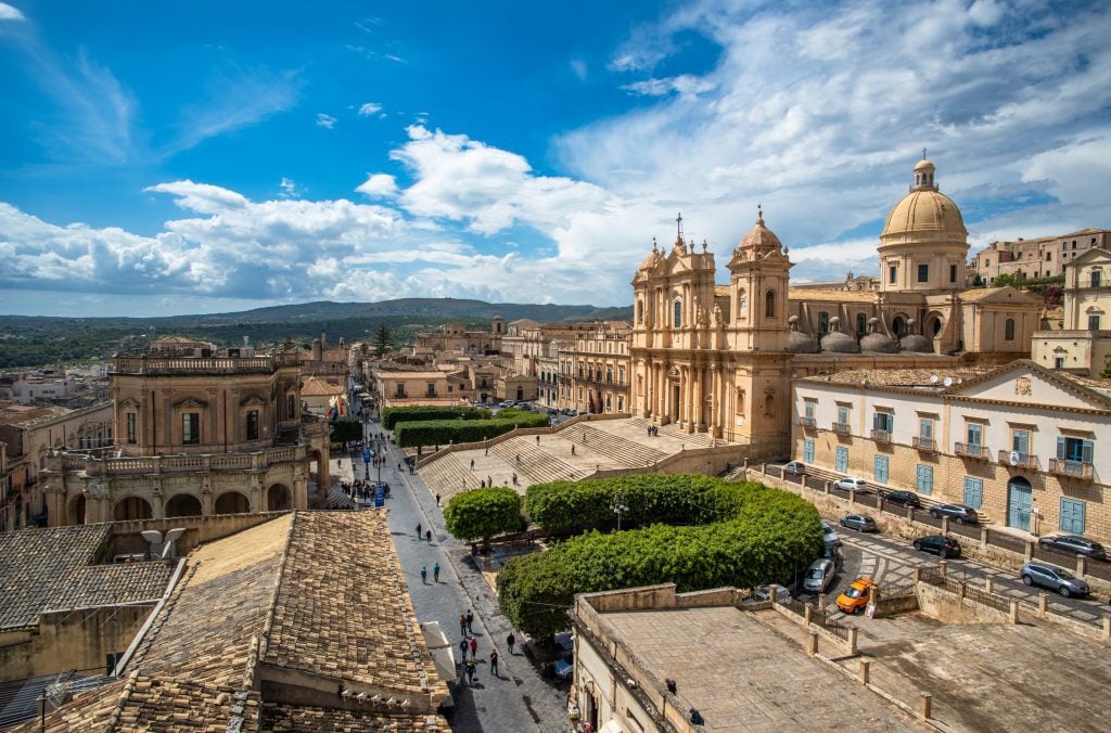 The city of Noto in Sicily, with a huge baroque cathedral with stairs leading up to it, and sand-colored stone buildings off into the distance.