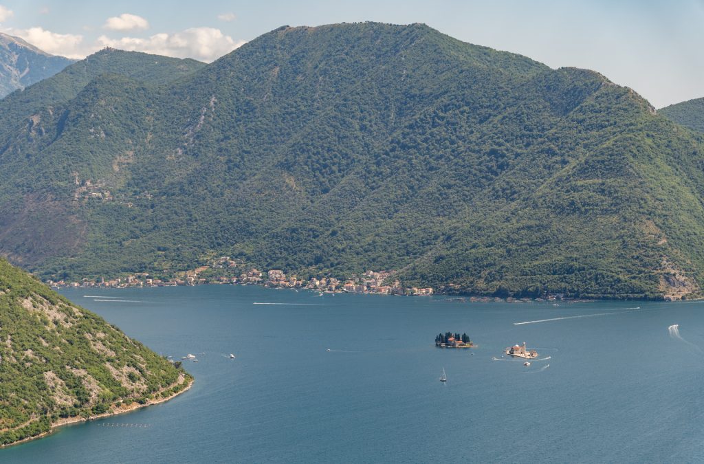 The Bay of Kotor from above, green mountains plunging into a deep blue fjord, two tiny islands in the water.