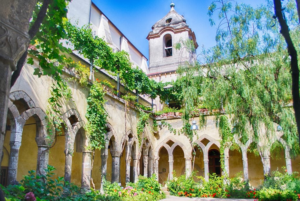 An open courtyard with pointy gothic-type windows, and plants growing wildly around them.