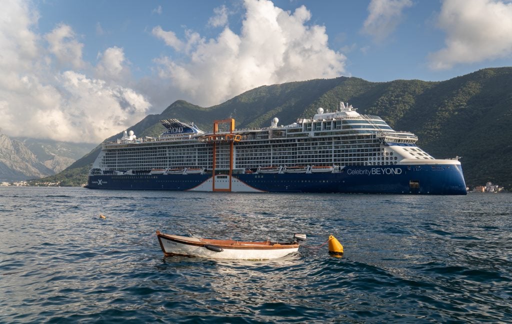 A giant cruise ship sailing through the Bay of Kotor, a tiny rowboat in the foreground.