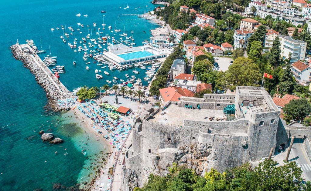 An overhead view of Herceg Novi's stone buildings in the old town, leading to the bright blue sea with lots of boats and a swimming pool right in the water.
