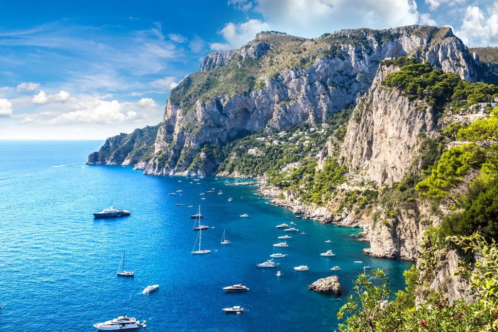 The rocky coast of Capri island, with dozens of small wooden boats in the bright blue water.