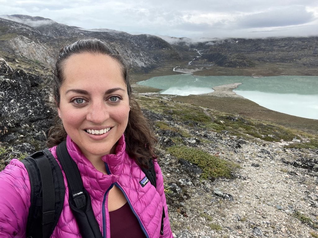 Kate taking a selfie wearing a pink jacket and hiking near a still green lake in Greenland.