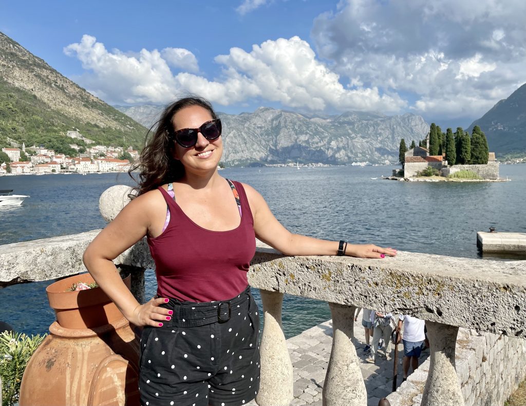 Kate standing in Perast in front of tiny St. George island, wearing a burgundy tank top and black polka dot shorts.