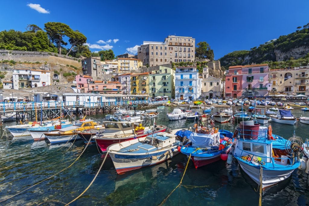 A marina full of colorful wooden rowboats; in the background are tall cliffs and colorful buildings on shore.