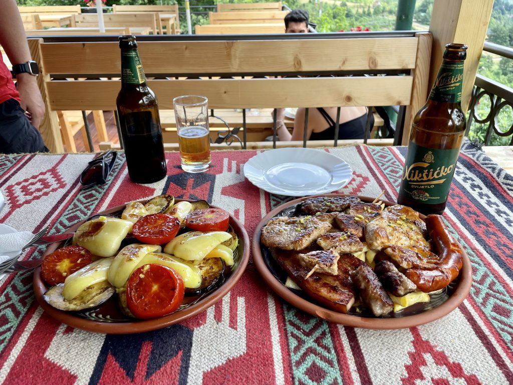 Two dishes on a patterned tablecloth in Montenegro: a plate of various grilled meats, and a plate or grilled tomatoes, peppers, and zucchini.